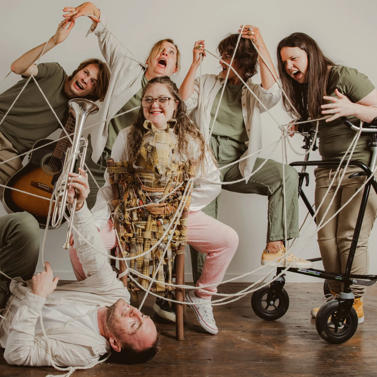 Six people pose playfully tangled in white rope, with a seated woman holding a trumpet at the centre and others gathered around her.