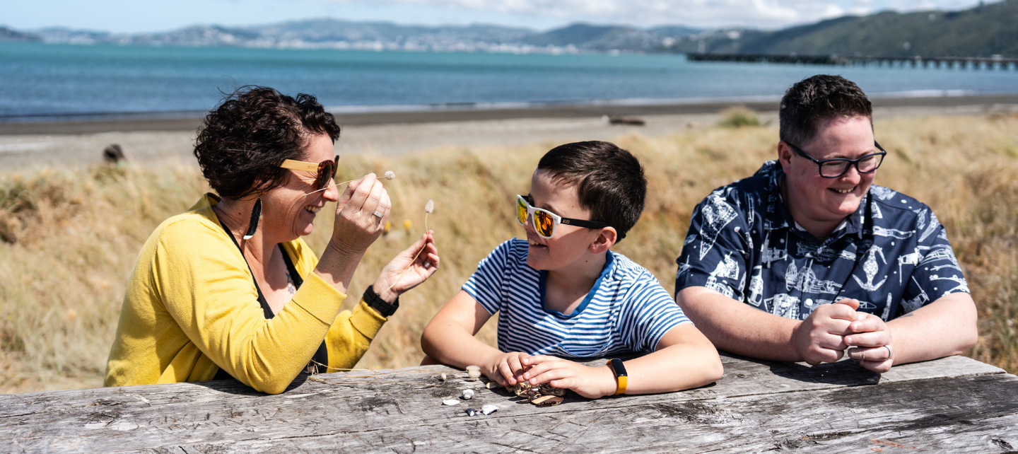 Three people sitting at a wooden bench at the beach. There is a young boy in the the middle smiling.