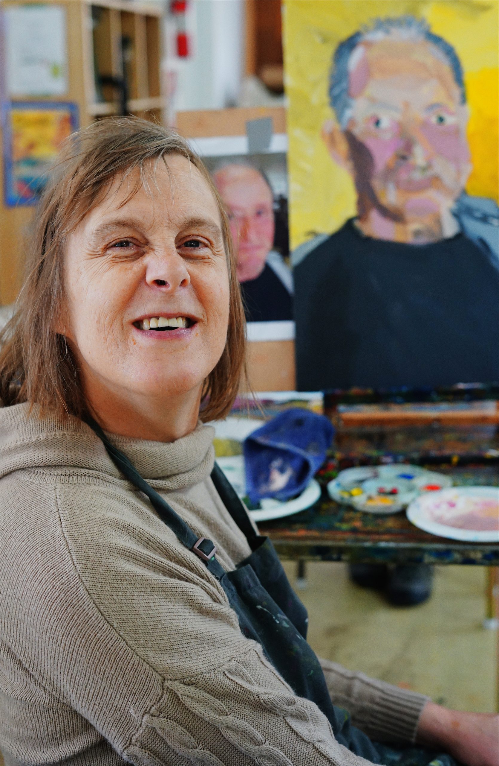 Woman sits in front of a painted portrait of a man, smiling.